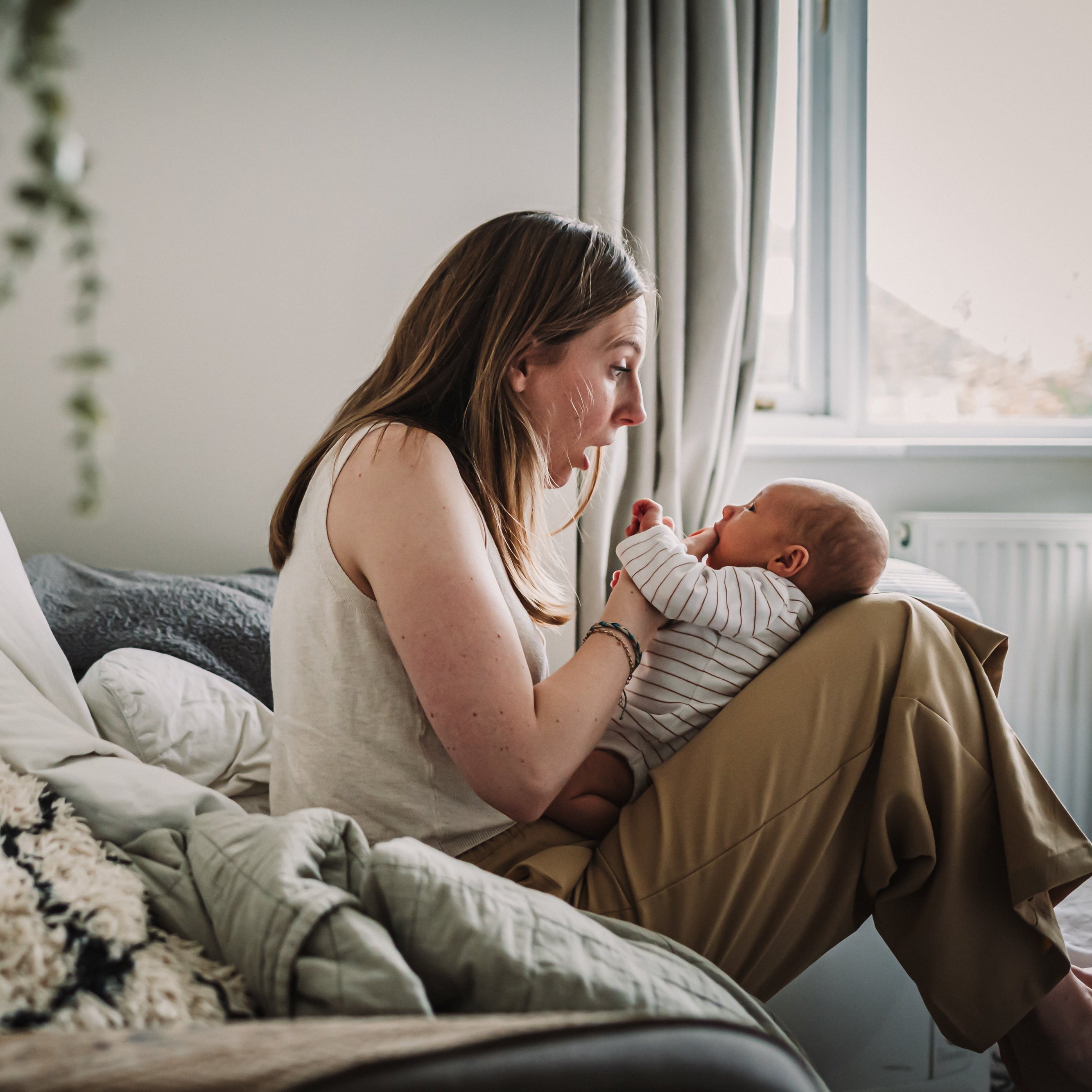 Woman holding a baby in a cozy room with a window and plant.
