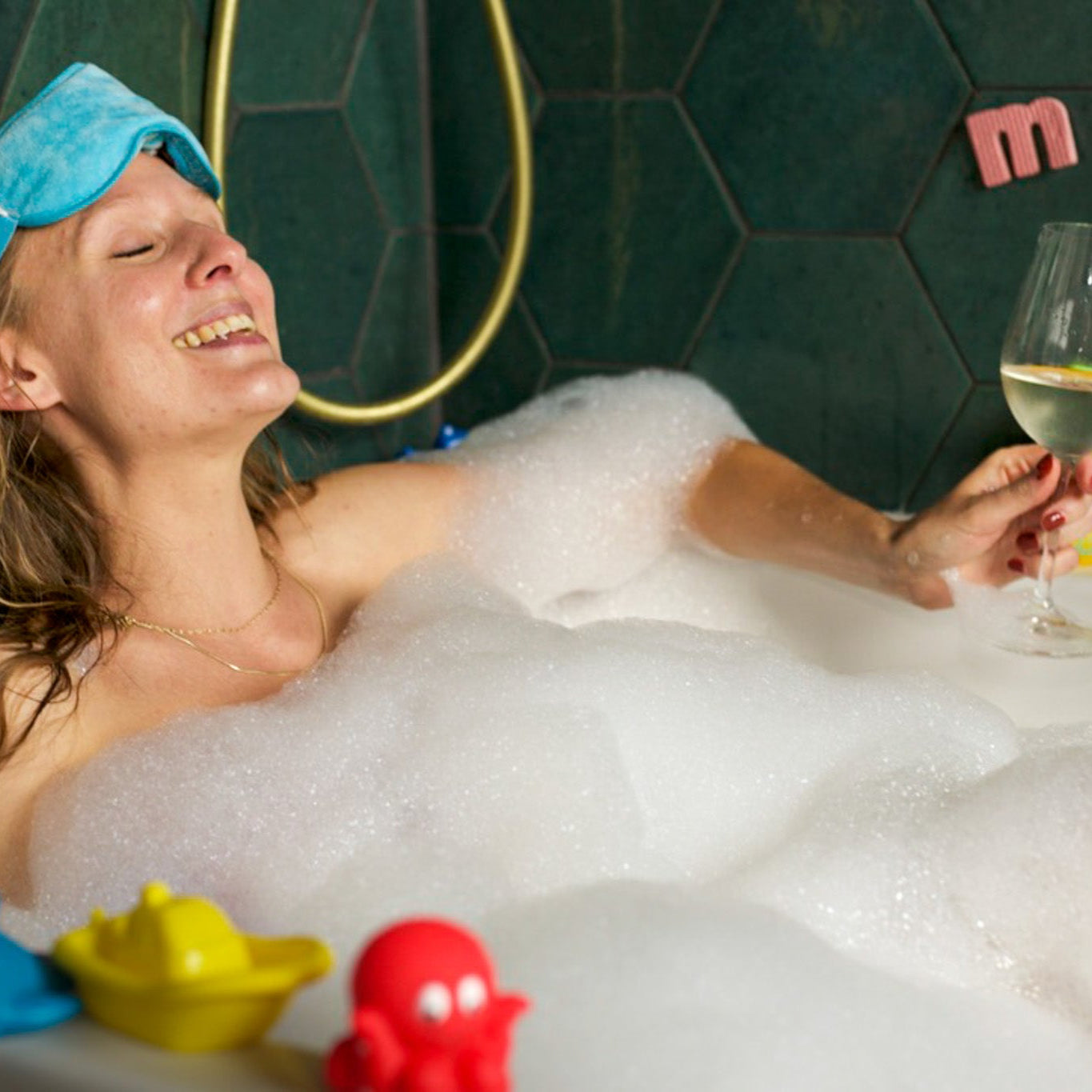 Woman in a bubble bath with toys and a glass of wine, wearing a sleep mask.